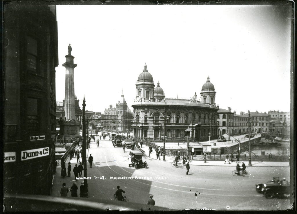 Black and white photo taken in 1922 at the junction of Whitefriargate and Princes Dock Street looking towards the Maritime Museum and Queen Victoria Square. The docks press up against the Maritime Museum. The dock has now been filled in and is now Queen's Gardens. People and cars are in the street leading towards and Queen Victoria Square.