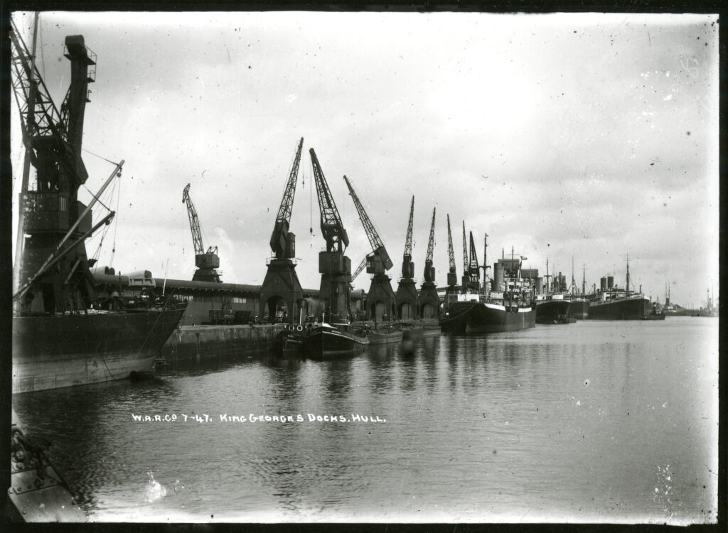 Black and white photo taken in 1922 of King George's Docks. The dock stretches from the bottom left to the top right of the frame with industrial cranes pointing skyward. Boats are moored against the dock.