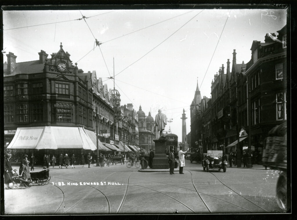 Black and white photo taken in 1922 at the junction of King Edward Street and Jameson Street, looking towards Queen Victoria Square. The pavements are busy with people shopping and all the shops lining King Edward's Street have canopies covering the pavements. A car drives around the statue in the middle of the junction towards the camera. 